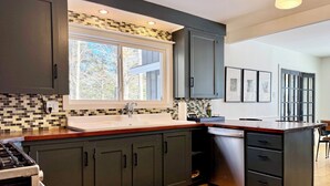 Une cuisine moderne avec des meubles de couleur sombre, un plan de travail en bois, un lave-vaisselle en acier inoxydable et une fenêtre donnant sur les arbres.