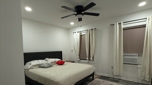 A bedroom with a bed, a ceiling fan, an air conditioning unit, and a patterned rug.
