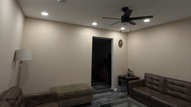 A modern living room with leather sofas, a glass coffee table, recessed ceiling lights, and a ceiling fan.
