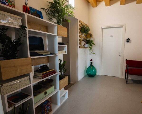 A room with white walls, a wooden floor, and a white door. There are shelves with various items, including books, a camera, and a green vase. - Panormos 91 Rooms & Terrace (Palermo)