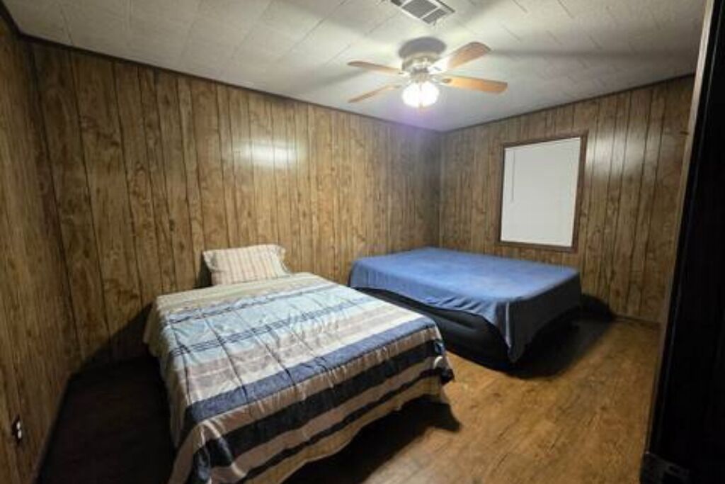 A room with wooden panel walls, a ceiling fan, a bed with a plaid bedspread, and a small table with a blue tablecloth.