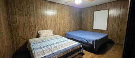A room with wooden panel walls, a ceiling fan, a bed with a plaid bedspread, and a small table with a blue tablecloth.