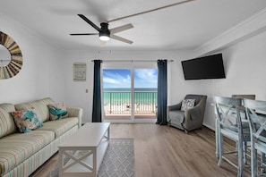 Una sala de estar con sofá, una mesa de centro, televisor montado en la pared y un balcón con vista a la playa.