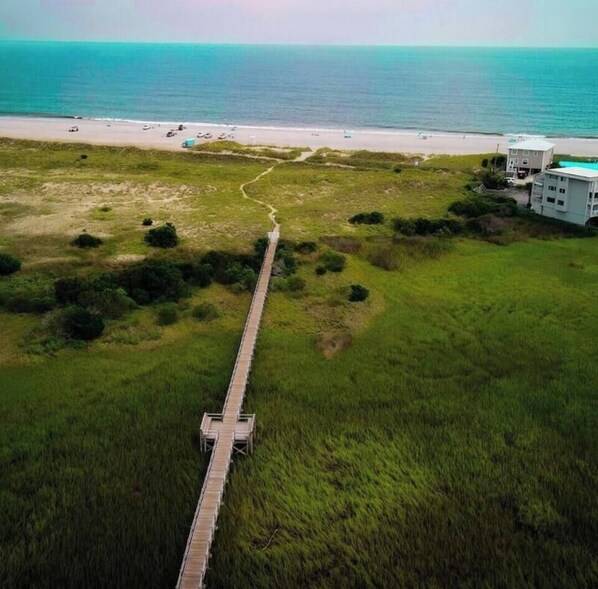 Eine Vogelperspektive eines Küstenabschnitts mit einer Holzpromenade, die zu einem Strand führt, umgeben von viel Grün und einem Gebäude mit weißer Fassade.