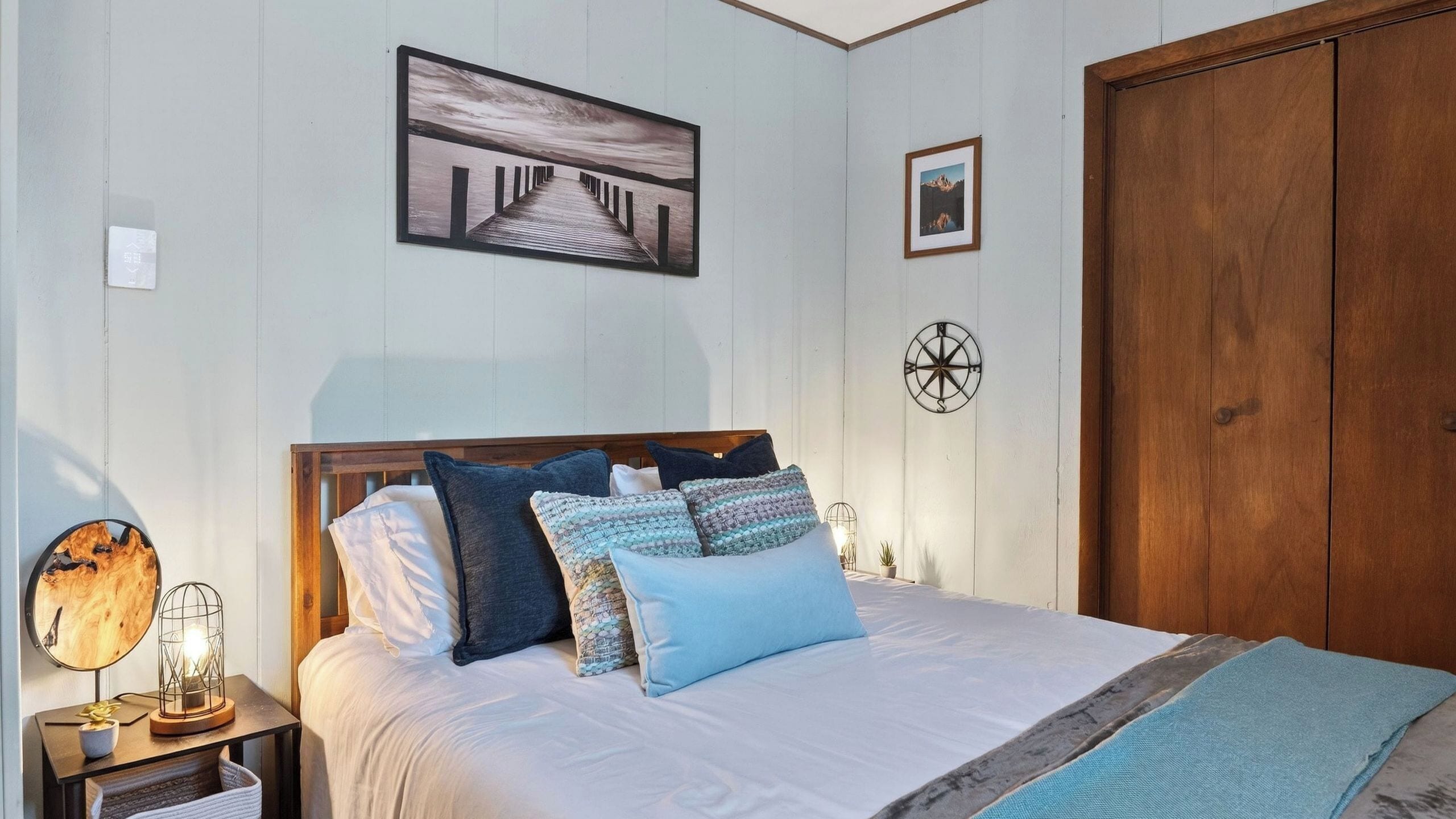 Een slaapkamer met een houten hoofdeinde, een bed met meerdere kussens, een nachtkastje met een lamp, een houten kledingkast en een aan de muur bevestigd kompas.