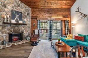 Ein gemütliches Blockhaus-Interieur mit Kamin aus Stein, Ledersessel, smaragdgrünem Sofa und einem 'Welcome'-Schild.