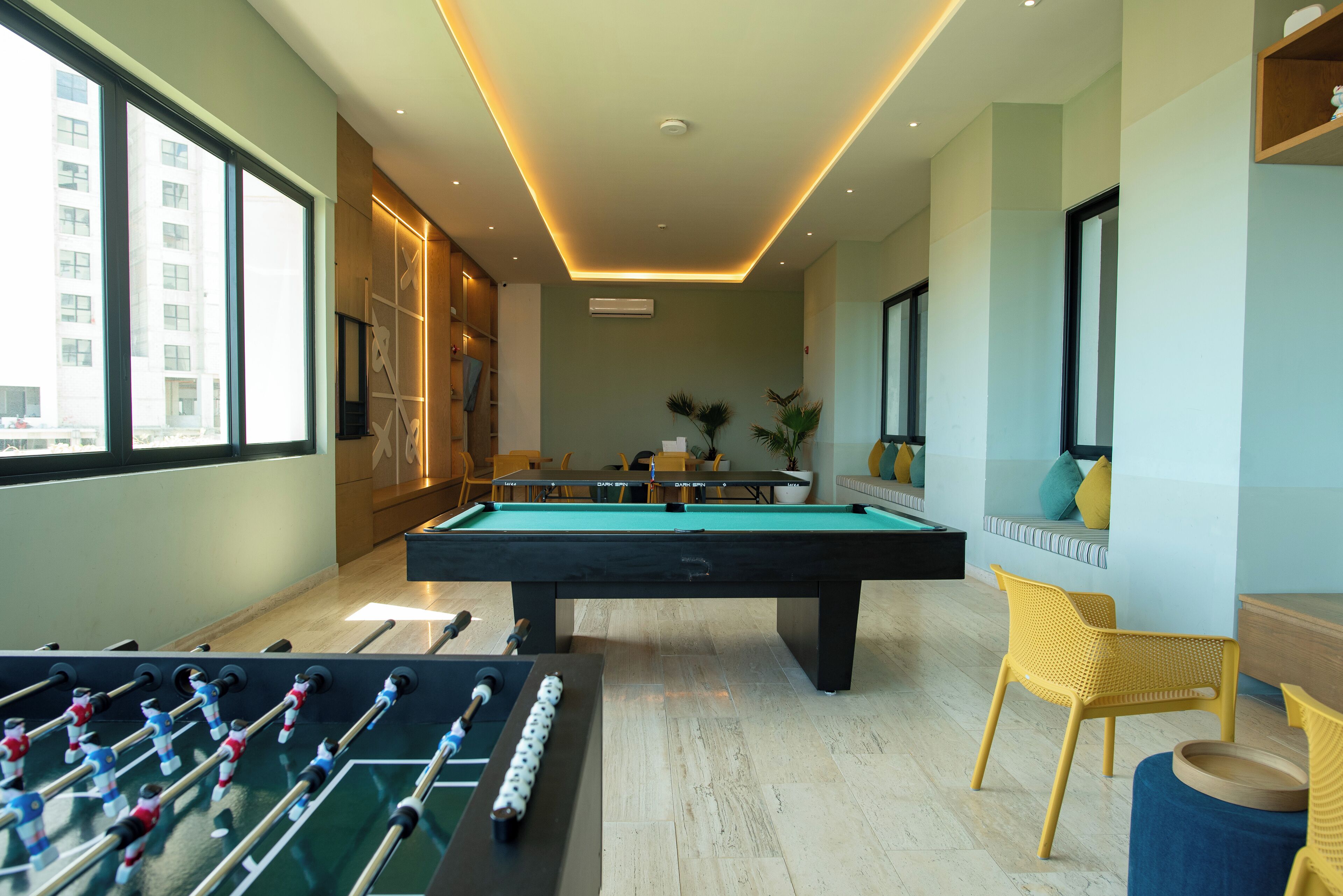 A pool table in a room with a foosball table, large windows, and a view of a building outside.