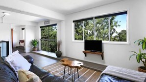 A modern living room with a sofa, a coffee table, and a TV mounted on the wall. There is a large window providing a view of trees outside. - 3 Bedrooms Vista Point Sanctuary (Burleigh Heads)