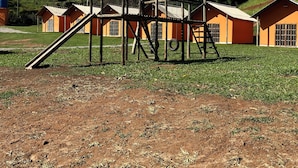 Un terrain de jeux avec un toboggan et une structure d’escalade, situé devant une rangée de bâtiments orange, avec un champ d’herbe et des collines en arrière-plan.