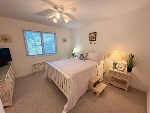 Um quarto com cama, ventilador de teto, janela, mesa de cabeceira com abajur e televisão.