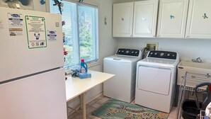 A compact kitchen with a refrigerator, washing machine, and oven. There is a window with blinds, a sink, and a countertop with various items. - Beach Front. Water Views. Inground Pool. (Cape May)