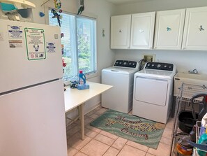 A compact kitchen with a refrigerator, washing machine, and oven. There is a window with blinds, a sink, and a countertop with various items.