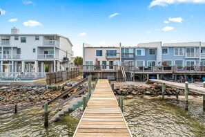 A wooden pier leading to a beachfront property with multiple-story buildings, a pool, and a rocky shoreline.