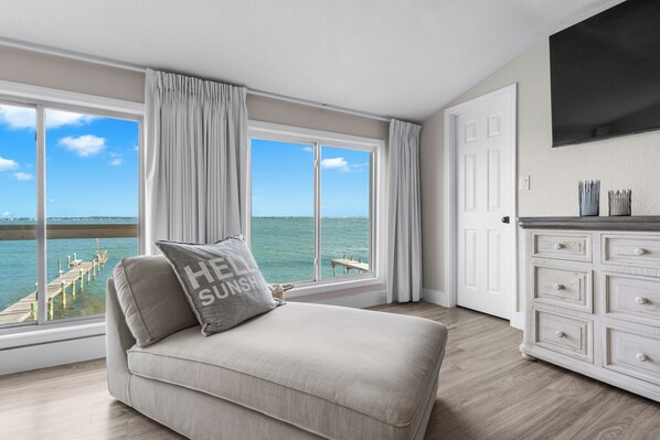 A room with a large window, a sofa with a pillow, a white dresser, and a flat-screen TV.