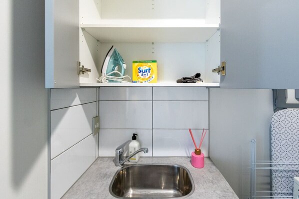 A kitchen with a stainless steel sink, a white shelf with a box of Sunlight dish soap, a white bottle of hand soap, and a pink soap dispenser. - 6BR Home | Walk to Wolfbrook Arena & Hagley Park (Christchurch)