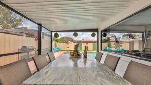 A covered outdoor dining area with a marble table, chairs, and a view of a backyard with a pool and a fence. - Versace Vibes Retreat, Pool Sauna Near Hard Rock (Hollywood)