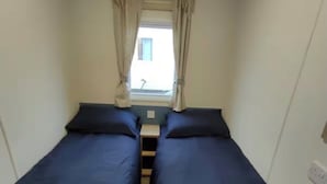 Ein schmales Zimmer mit zwei Einzelbetten, einem fenster mit Vorhängen und einem kleinen Tisch in der Mitte.