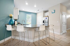 A modern kitchen with a central island, white bar stools, a blue vase with flowers, and a refrigerator. - 20 min to Disney , Epic , Star Wars , Gameroom ,Pool Luxe dw7 (Davenport)