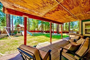 Ein überdachter Außenbereich mit Holzboden, Sitzgelegenheiten und Blick auf einen Garten mit Schaukel und Bäumen.