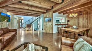 Un vaste espace de vie agrémenté de poutres en bois, un escalier, un coin repas avec une table et des chaises en bois, et une grande fenêtre offrant une vue sur les montagnes.