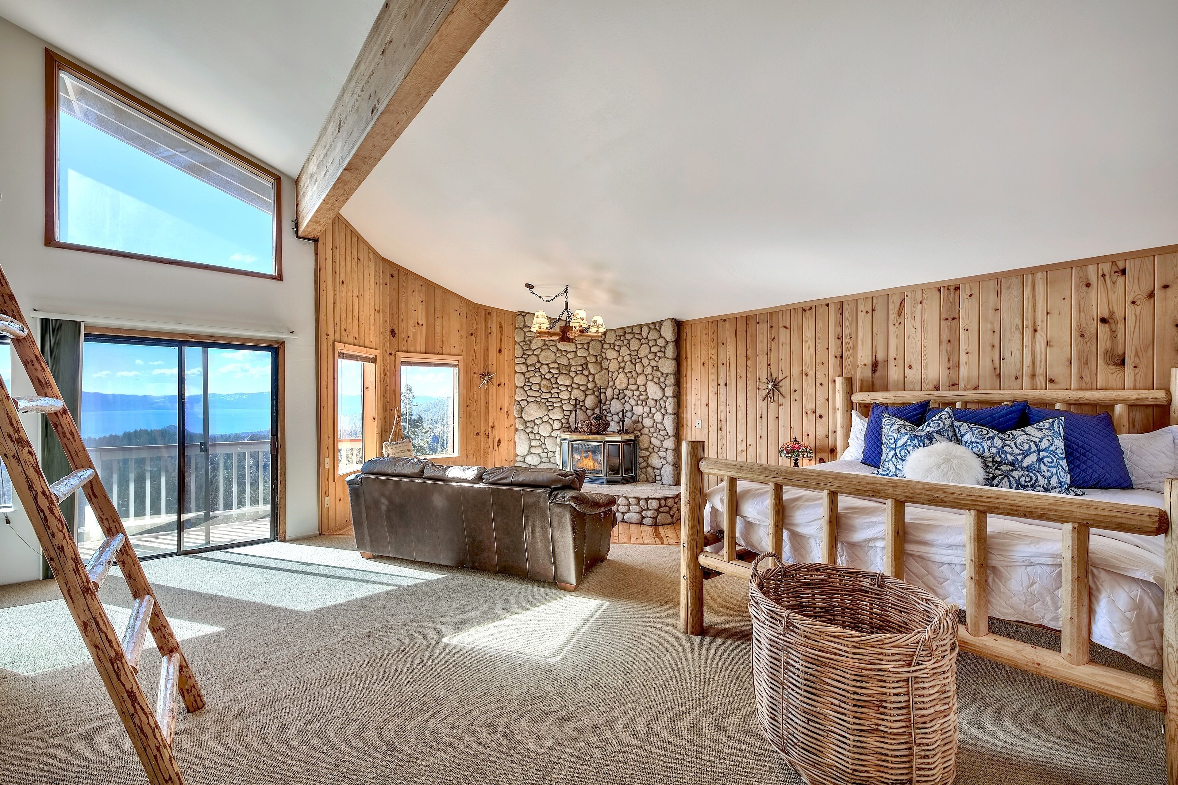 A spacious room with a loft bed, a stone fireplace, a wooden ceiling, and a large window overlooking a balcony.