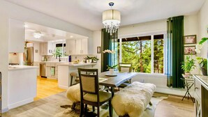 A modern kitchen with white cabinets, a dining area with a wooden table and chairs, a chandelier, and a view of greenery outside. - LX35  The Wolf Round Hill Retreat in the heart of Lake Tahoe (Zephyr Cove)