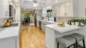 A modern kitchen with white cabinetry, a central island, a dining area with a table and chairs, and a view of greenery outside. - LX35  The Wolf Round Hill Retreat in the heart of Lake Tahoe (Zephyr Cove)