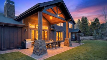 Una casa de dos pisos con una chimenea de piedra, ventanas amplias y un porche cubierto con mobiliario exterior.