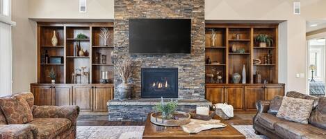 A living room with a stone fireplace, wooden shelves, and a large flat-screen TV.