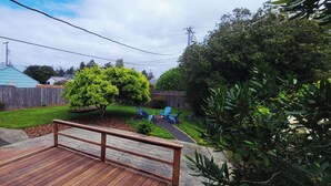 Un patio trasero con terraza de madera, zona cercada, un árbol y un pequeño jardín con sillas azules.
