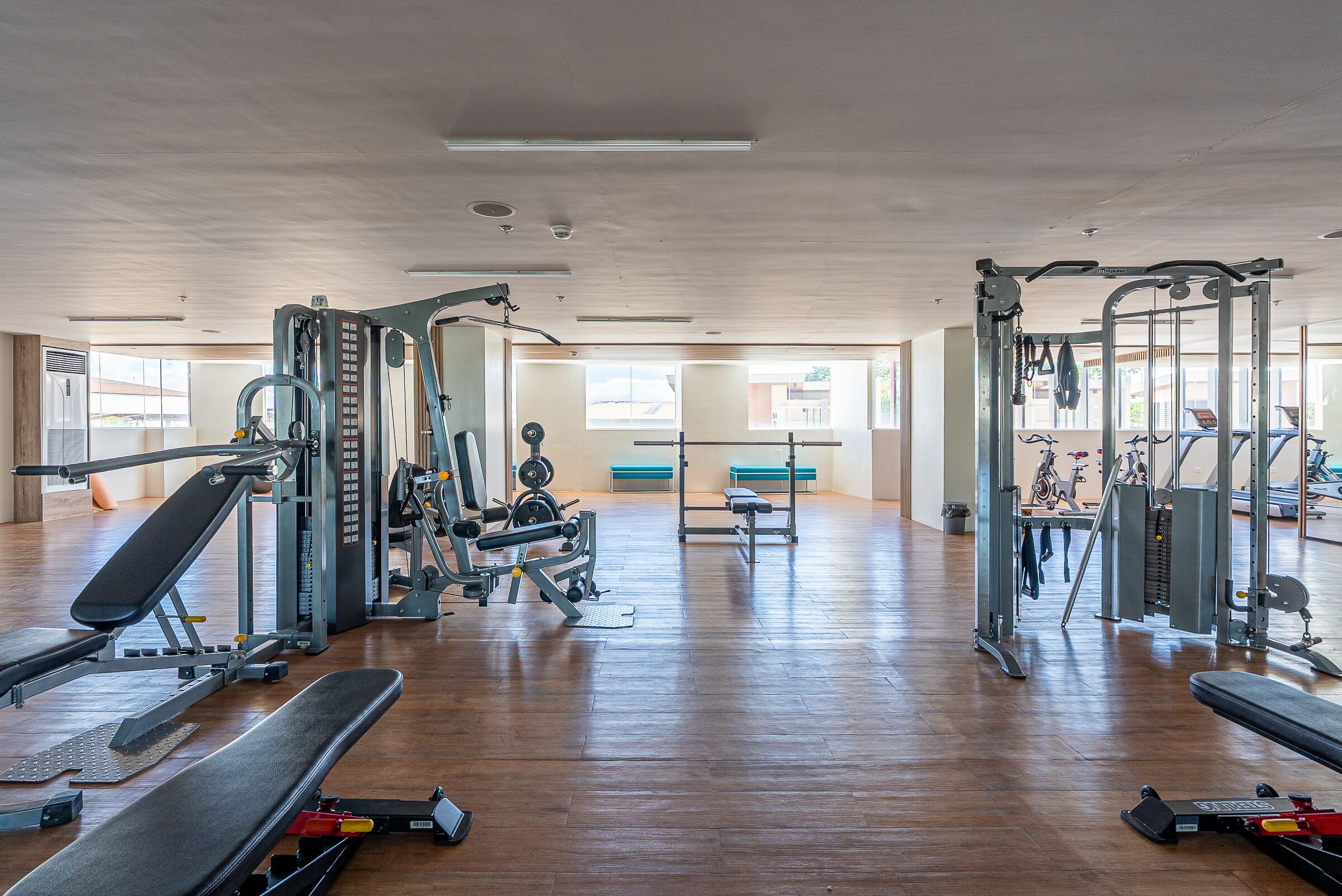 Un gimnasio con diverso equipo de ejercicio, pisos de madera y ventanas amplias.