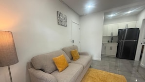 A modern living room with a grey sofa, two mustard-colored cushions, a floor lamp, and a kitchen area with white cabinets and a black refrigerator. - The Warm Stay  4 (Miami)