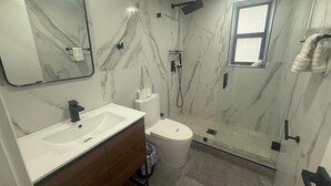 A modern bathroom with a marble wall, a sink with a wooden vanity, a toilet, and a shower area. - The Warm Stay  4 (Miami)