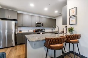 A modern kitchen with grey cabinets, stainless steel appliances, and a granite countertop. There are two bar stools with leather seats. - Designer Apt: Walk to Busch Stadium, Free Parking (St. Louis)