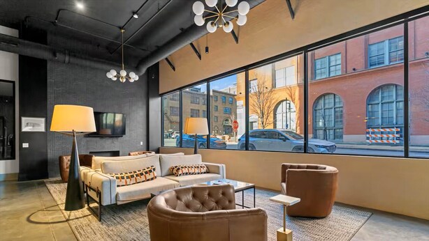 A modern lounge area with a sofa set, a leather armchair, and a coffee table. There are two lamps, a flat-screen TV mounted on the wall, and a large window offering a view of the street. - Designer Apt: Walk to Busch Stadium, Free Parking (St. Louis)