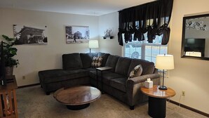 A living room with a sectional sofa, a round coffee table, a side table with a lamp, and two wall-mounted paintings. - Whole House - Sleeps 6 - Remodeled Kitchen & Bath - Near I-90 - North Side Jvl (Janesville)