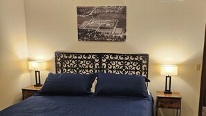A neatly made bed with a dark blue bedspread and two pillows, flanked by two bedside tables with lamps, in a room with a simple, modern design. - Whole House - Sleeps 6 - Remodeled Kitchen & Bath - Near I-90 - North Side Jvl (Janesville)