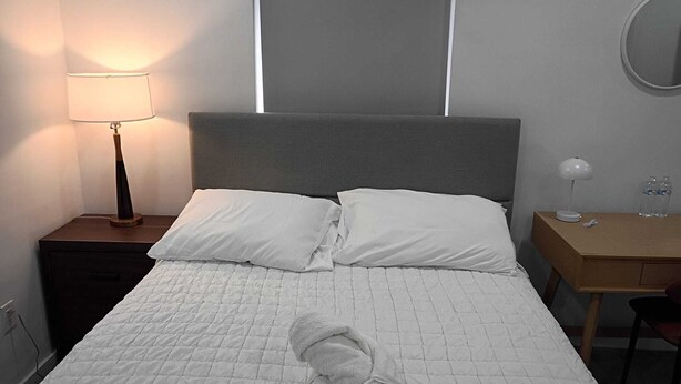 Cama bem arrumada com duas almofadas, mesa de cabeceira com abajur e pequena escrivaninha com espelho e garrafa de água.