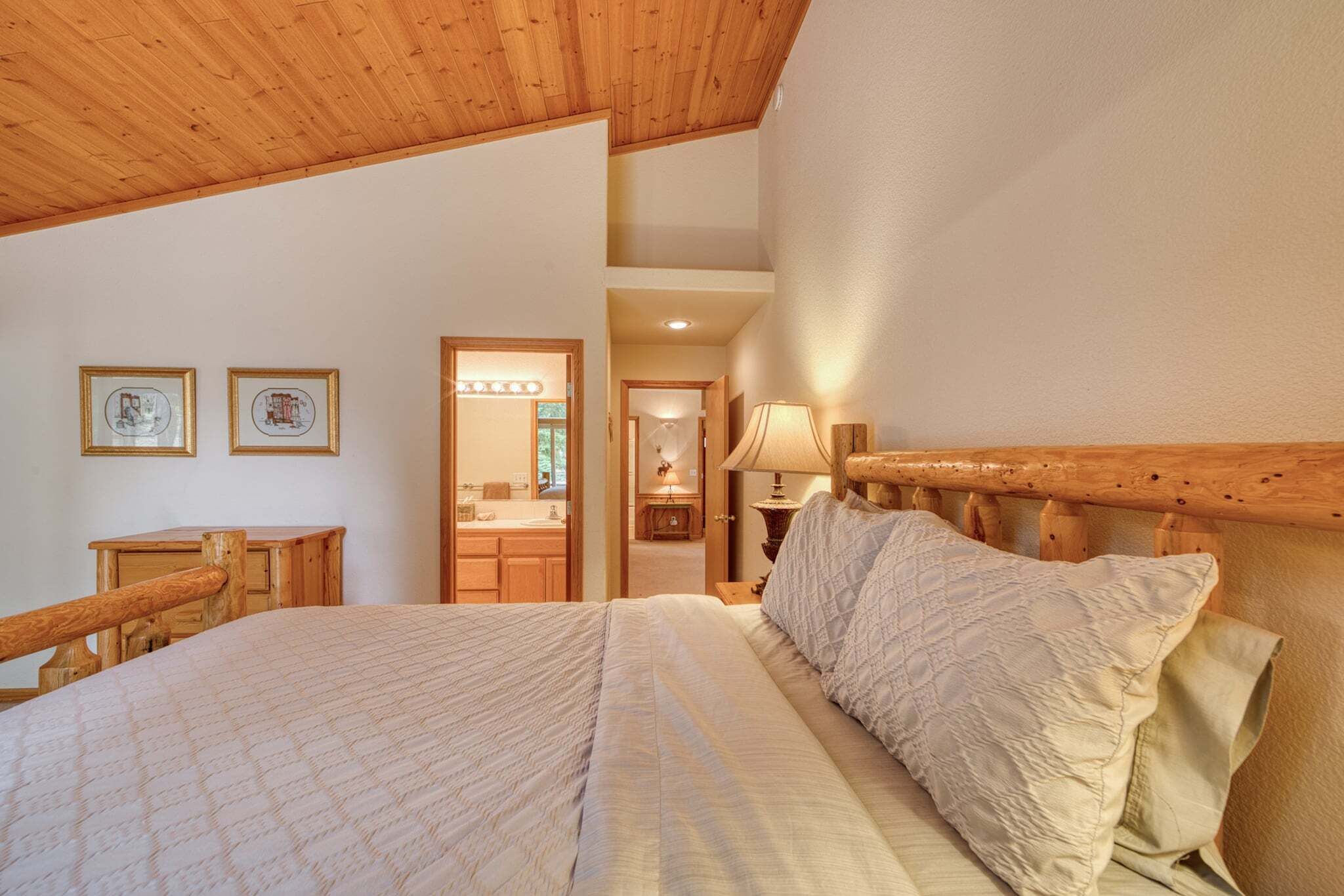 A bedroom with a wooden headboard, a bed with a quilted bedspread, and a nightstand with a lamp. There is a wooden dresser and a framed picture on the wall.