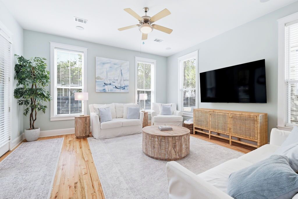 A living room with a ceiling fan, white sofas, a round coffee table, a flat-screen TV, and a large window with blinds.