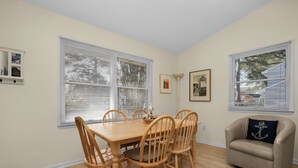 Un comedor con una mesa y sillas de madera, un sillón color crema y dos ventanas con persianas.
