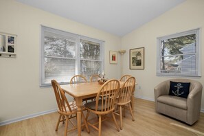 A dining area with a wooden table and chairs, a beige armchair, and two windows with blinds. - Half Shell | Clean accommodations close to Beach Road! (Chincoteague Island)