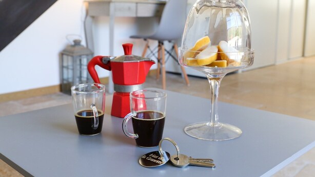 A table with a coffee maker, two cups of coffee, a glass with lemon slices, and a set of keys.