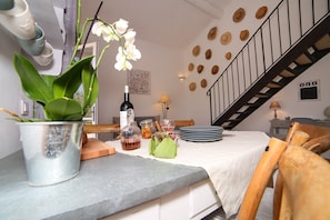 Ein Essbereich mit Tisch für zwei Personen, einem Topf mit Orchideen, einer Weinflasche und einer Obstschale. Im Hintergrund ist eine Treppe zu sehen.