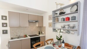 Een kleine keuken met een spoelbak, fornuis en kastjes, een eettafel met stoelen en een plank met keukengerei.