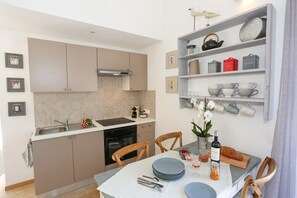A compact kitchen with a sink, stove, and cabinets, a dining table with chairs, and a shelf with kitchenware.