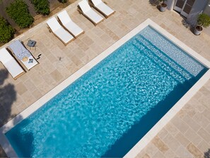 A rectangular swimming pool with clear blue water, surrounded by beige tiles. There are four white lounge chairs aligned along one side of the pool.
