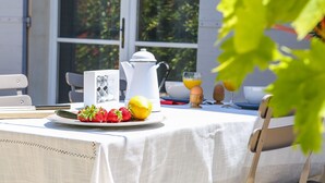 Una mesa de desayuno con mantel blanco, un plato de fruta, una tetera blanca y un vaso de jugo de naranja.