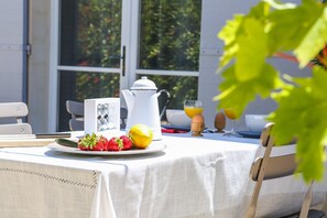 Una mesa de desayuno con mantel blanco, un plato de fruta, una tetera blanca y un vaso de jugo de naranja.