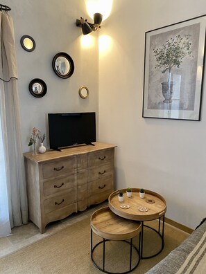 A room with a flat-screen TV on a wooden dresser, a round wooden table with two small potted plants, and a framed artwork on the wall.